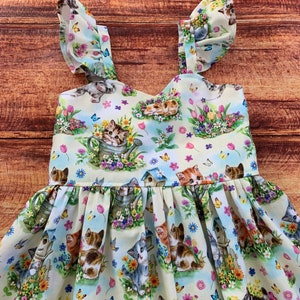 K&ouml;nnte beinhalten: Ein &auml;rmelloses Kleid aus hellem Stoff mit einem wiederkehrenden Muster aus K&auml;tzchen, Vogelh&auml;uschen, bunten Tulpen, G&auml;nsebl&uuml;mchen und Schmetterlingen. Das Oberteil hat ger&uuml;schte Tr&auml;ger und einen gerafften Rock mit einer vollen, plissierten Form.