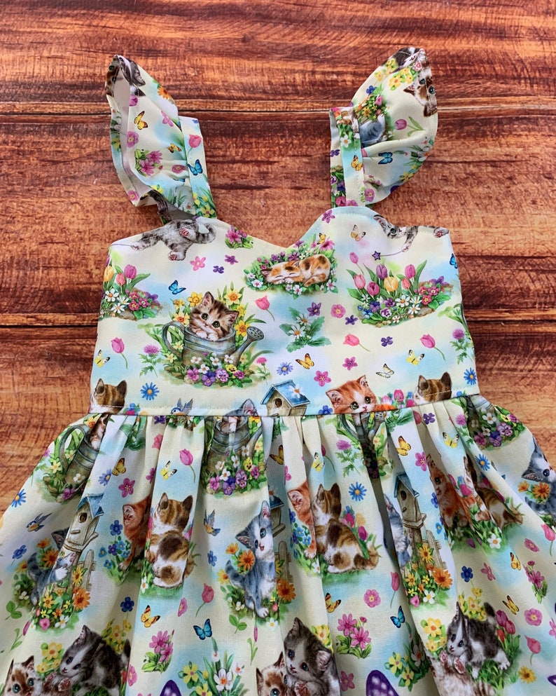 K&ouml;nnte beinhalten: Ein &auml;rmelloses Kleid aus hellem Stoff mit einem wiederkehrenden Muster aus K&auml;tzchen, Vogelh&auml;uschen, bunten Tulpen, G&auml;nsebl&uuml;mchen und Schmetterlingen. Das Oberteil hat ger&uuml;schte Tr&auml;ger und einen gerafften Rock mit einer vollen, plissierten Form.