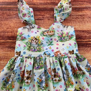 K&ouml;nnte beinhalten: Ein &auml;rmelloses Kleid mit hellblauem Hintergrund und einem wiederkehrenden Muster aus K&auml;tzchen, bunten Blumen, Schmetterlingen und Vogelh&auml;uschen. Das Oberteil hat einen herzf&ouml;rmigen Ausschnitt und die Tr&auml;ger sind mit R&uuml;schen versehen.