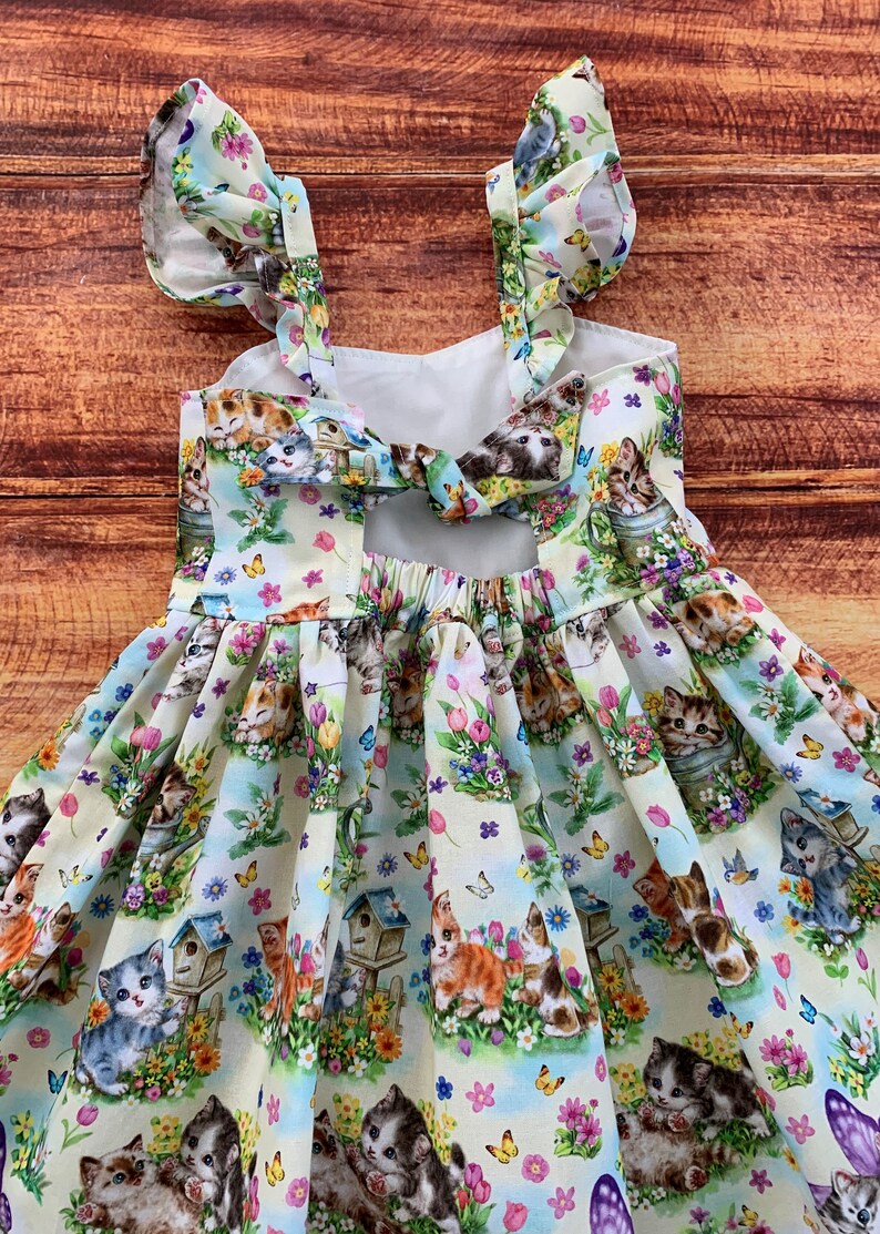 K&ouml;nnte beinhalten: Ein &auml;rmelloses Kleid mit cremefarbenem Hintergrund und einem bunten Druck aus K&auml;tzchen, Vogelh&auml;uschen, Blumen und Schmetterlingen. Das Oberteil hat ger&uuml;schte Tr&auml;ger und eine Bindedetail am R&uuml;cken, der Rock ist in der Taille gerafft.