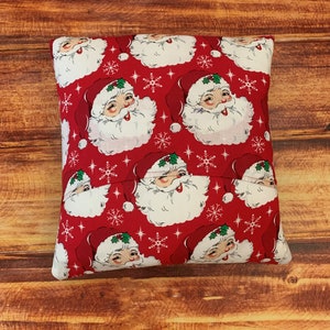K&ouml;nnte beinhalten: Ein quadratisches, rotes Weihnachtskissen mit einem sich wiederholenden Muster von Weihnachtsmann-Gesichtern und wei&szlig;en Schneeflocken. Der Weihnachtsmann tr&auml;gt einen roten Hut mit wei&szlig;em Rand und einen wei&szlig;en Bart. Das Kissen liegt auf einer Holzoberfl&auml;che.