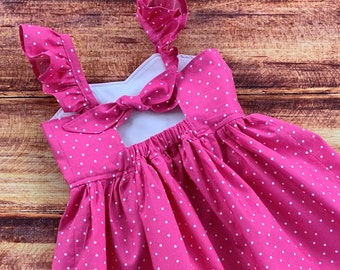 May include: A bright pink dress featuring a white polka dot pattern, ruffled shoulder straps, and a tie-back closure. The bodice includes a cutout detail, while the skirt is gathered at the waist with an elastic band, creating a full silhouette.