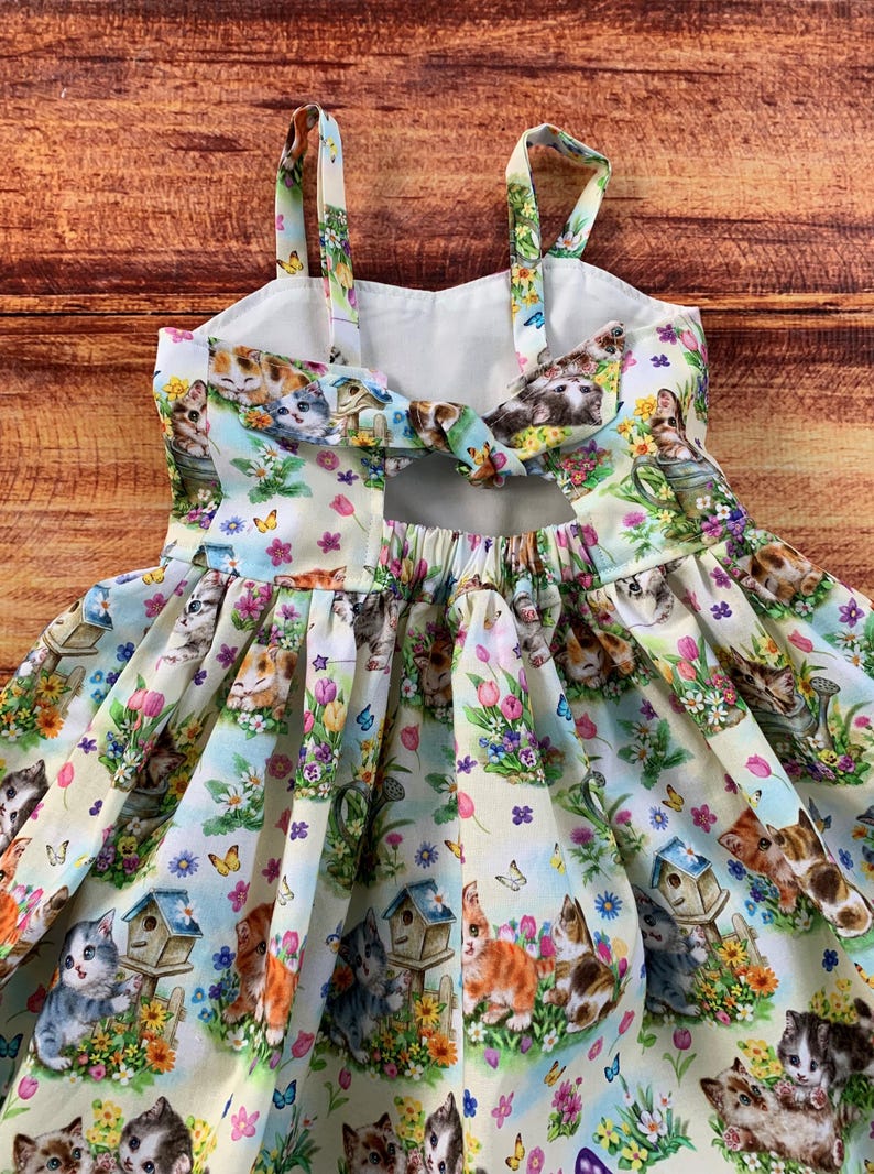 Pu&ograve; includere: Un abito senza maniche con sfondo color crema chiaro e un motivo ripetuto di gattini, casette per uccelli, fiori colorati e farfalle. Il corpetto presenta spalline sottili e un'apertura sulla schiena con dettaglio a fiocco.