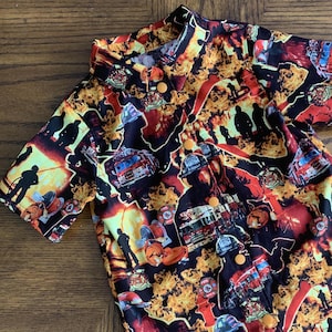 May include: A short-sleeved button-up shirt with a black background and a colorful print of firetrucks, firefighters, and flames. The shirt has a mandarin collar and yellow buttons.
