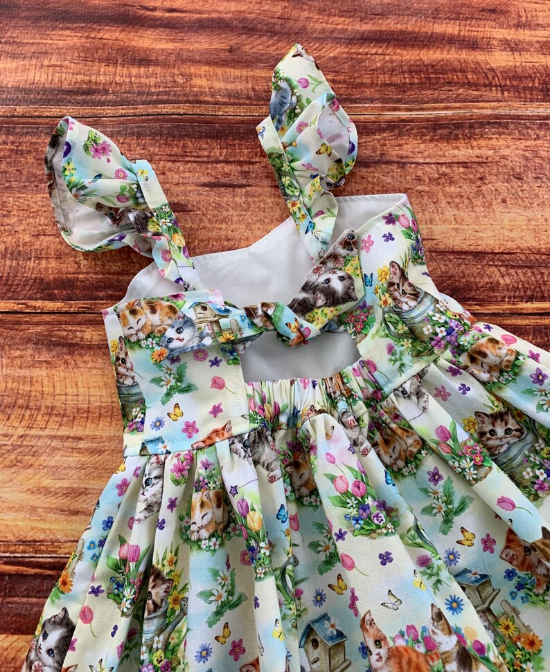 K&ouml;nnte beinhalten: Ein &auml;rmelloses Kleid aus hellem Stoff mit einem wiederkehrenden Muster aus K&auml;tzchen, bunten Fr&uuml;hlingsblumen, Schmetterlingen und Vogelh&auml;uschen. Das Kleid hat ger&uuml;schte Schultertr&auml;ger und einen Bindeg&uuml;rtel am oberen R&uuml;ckenbereich.