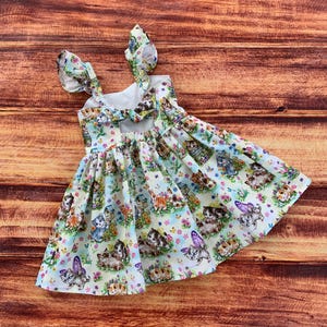 K&ouml;nnte beinhalten: Ein &auml;rmelloses Kleid mit hellblauem Hintergrund und einem sich wiederholenden Muster aus K&auml;tzchen, Schmetterlingen und Vogelh&auml;uschen. Das Kleid hat ger&uuml;schte Schultertr&auml;ger und eine Schleife am R&uuml;ckenausschnitt sowie einen gerafften Rock mit wei&szlig;em Futter.