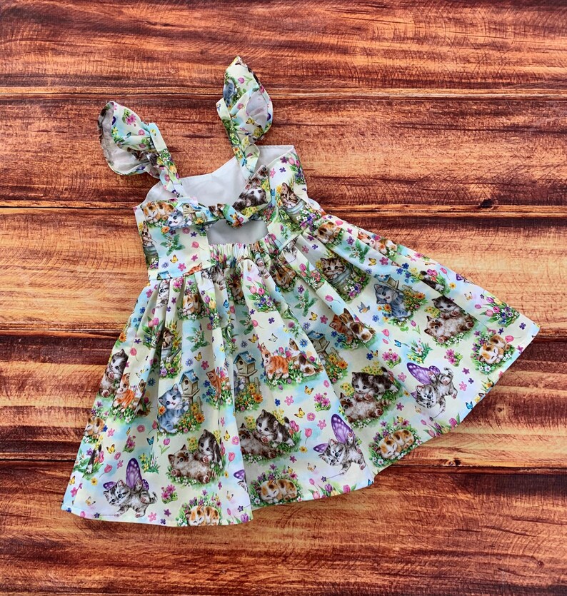 K&ouml;nnte beinhalten: Ein &auml;rmelloses Kleid mit hellblauem Hintergrund und einem sich wiederholenden Muster aus K&auml;tzchen, Schmetterlingen und Vogelh&auml;uschen. Das Kleid hat ger&uuml;schte Schultertr&auml;ger und eine Schleife am R&uuml;ckenausschnitt sowie einen gerafften Rock mit wei&szlig;em Futter.