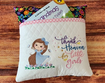 May include: White quilted pillow with an embroidered design featuring a cartoon girl in a blue dress, butterflies, and the text "thank Heaven for little girls." The pillow has a pocket holding a children's book.