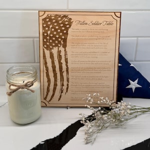 May include: A wooden plaque with an engraved American flag and text, "Fallen Soldier Table." A lit candle in a mason jar with a twine bow sits to the left. A folded American flag and flowers are in the background.