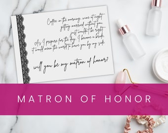 May include: A white card with black lace trim and black calligraphy text that reads "Coffee in the morning, wine at night getting married without you. Just wouldn't be right. As I prepare for the day, I become a bride. It would mean the world to have you by my side. Will you be my matron of honor?" The card is on a white marble surface with a pink and gold face roller, a white jar with white cream, and two clear glass bottles with rose gold lids.