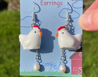 Pode incluir: Brincos Chicken and the Egg. Brincos brancos em forma de galinha com cristas vermelhas e bicos laranja. Cada brinco tem uma pérola pendurada. Os brincos são exibidos em um cartão azul com o texto "Chicken and the Egg Earrings".