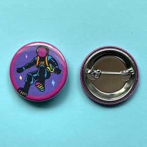 May include: A pink and purple button with a cartoon astronaut wearing a black suit and rainbow socks. The astronaut is floating in space with stars around them. The back of the button is silver with a pin.