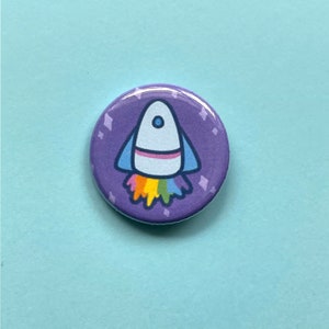 May include: A purple button with a white rocket ship with a rainbow flame. The rocket ship is on a purple background with white stars.