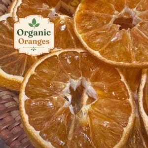 May include: Several dehydrated orange slices with a translucent, deep amber texture. A white rectangular label with a decorative border is overlaid on the fruit, featuring a green leaf icon and the text Organic Oranges in green and orange.