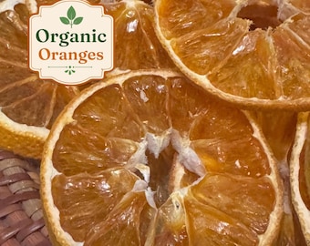 May include: Several dehydrated orange slices with a translucent, deep amber texture. A white rectangular label with a decorative border is overlaid on the fruit, featuring a green leaf icon and the text Organic Oranges in green and orange.