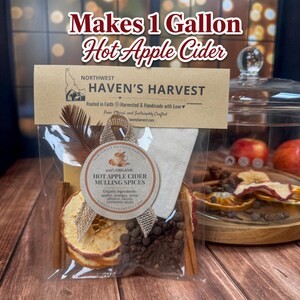 May include: A clear plastic pouch containing hot apple cider mulling spices, including dried apple slices, cinnamon sticks, and whole cloves. The label reads Makes 1 Gallon Hot Apple Cider and Northwest Haven's Harvest, with a round tag listing organic ingredients.