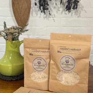 K&ouml;nnte beinhalten: Drei braune Papiert&uuml;ten mit Sweet Lilac Infused Sugar von Northwest Haven's Harvest. Die T&uuml;ten haben ein klares Fenster, das den Zucker zeigt. Die T&uuml;ten stehen auf einer Holzoberfl&auml;che. Der Hintergrund umfasst eine gr&uuml;ne Vase, Trockenblumen und ein Holzschneidebrett.