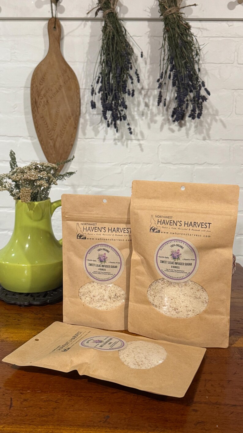 K&ouml;nnte beinhalten: Drei braune Papiert&uuml;ten mit Sweet Lilac Infused Sugar von Northwest Haven's Harvest. Die T&uuml;ten haben ein klares Fenster, das den Zucker zeigt. Die T&uuml;ten stehen auf einer Holzoberfl&auml;che. Der Hintergrund umfasst eine gr&uuml;ne Vase, Trockenblumen und ein Holzschneidebrett.