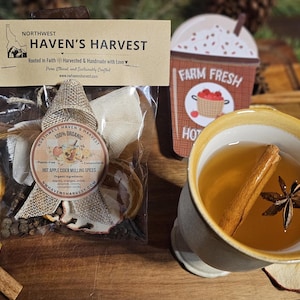 May include: A clear bag of Northwest Haven's Harvest Hot Apple Cider Mulling Spices with a decorative bow. A mug of hot cider with a cinnamon stick and star anise is next to a Farm Fresh sign. Dried orange slices and cinnamon sticks are also visible.