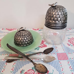 May include: A glass jar with a silver-colored beehive-shaped lid and a small bee on top. A matching lid sits on a green plate. Three antique spoons are crossed on a floral patterned tablecloth.