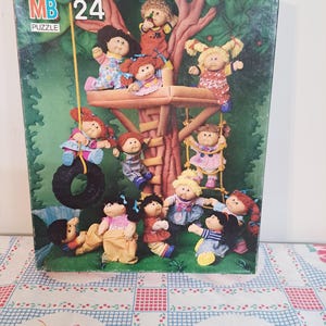 May include: A rectangular cardboard puzzle box featuring a colorful illustration of eleven Cabbage Patch Kids dolls playing in and around a large treehouse. The box displays the MB Puzzle logo and the number 24 in the upper left corner.