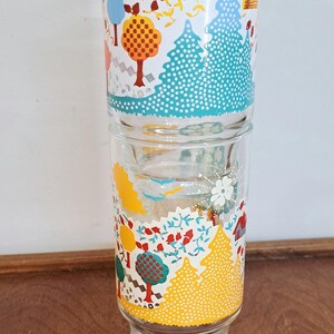 May include: Two clear drinking glasses with a colorful, whimsical forest design. The glasses feature trees, flowers, and houses in shades of yellow, blue, red, and white. The glasses are stacked on top of each other.