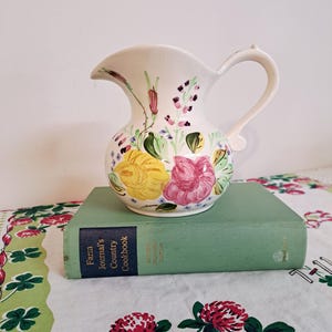 May include: A cream-colored ceramic pitcher with a handle, adorned with hand-painted flowers in yellow, pink, and purple. The pitcher is resting on a vintage green book, adding a touch of rustic charm. The pitcher is approximately 8 inches tall.
