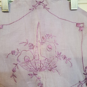 May include: A lavender-colored textile with a detailed embroidered design. The embroidery features a basket of flowers in shades of purple, surrounded by delicate floral accents. The textile is hanging from a silver hanger.