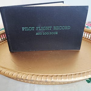 May include: A black Pilot Flight Record and Log Book with green lettering rests on a gold-colored, decorative tray. The book is slightly open, revealing the white pages inside. The tray has a scalloped edge.