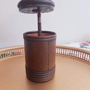 May include: A brown and black wooden container with a metal lid. The lid has a small hole in the center and a metal rod extending from the hole. The container is sitting on a gold tray.