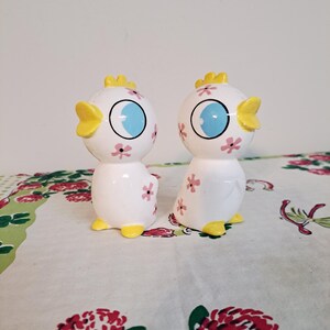 May include: A pair of white ceramic salt and pepper shakers shaped like cartoon birds. Each bird has a yellow crest, yellow feet, and a blue eye. Pink flower accents decorate the body. The shakers are on a floral tablecloth.