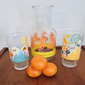 May include: A clear glass pitcher and two matching glasses with orange slice designs. The pitcher has a yellow band at the base. Three oranges sit in front of the glassware. The glasses have colorful landscape designs.