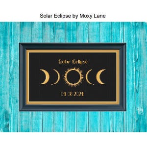 May include: A black and gold cross-stitch pattern featuring a solar eclipse with three crescent moons and the date "04 06 2024". The pattern is framed in a black frame with a gold trim and is displayed against a turquoise wooden background.