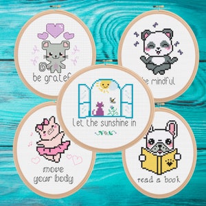 May include: Five cross-stitch embroidery hoops with cute designs. The designs include a mouse with the text "be grateful", a panda with "be mindful", a pig in a tutu with "move your body", a cat and window with "Let the sunshine in", and a dog reading a book.