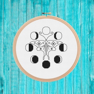 Può includere: Un ricamo in bianco e nero di una farfalla con otto fasi lunari che la circondano. Il disegno è ricamato su tessuto bianco e incorniciato in un cerchio di legno.