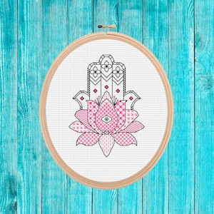 May include: A pink and black hamsa hand with an evil eye in the center, surrounded by a flower design, is displayed in a wooden embroidery hoop.