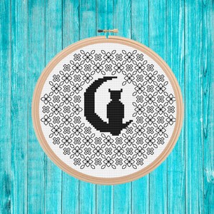 Può includere: Un telaio da ricamo a punto croce con un gatto nero su una luna crescente, circondato da un motivo floreale ripetuto in bianco e nero. Il telaio in legno ha un diametro di circa 15 cm, su uno sfondo di legno turchese.