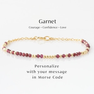 May include: A gold-colored bracelet with small, round, red garnet beads and gold beads. The text "Garnet" is above the bracelet, with "Courage • Confidence • Love" below it. The text "Personalize with your message in Morse Code" is below the bracelet.