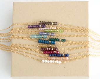 May include: A set of 14 gold necklaces with different colored gemstone beads. Each necklace features a bar of beads in a single color, including amethyst, rose quartz, peridot, aquamarine, blue topaz, emerald, citrine, garnet, sapphire, and moonstone.