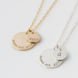 May include: Two gold and silver necklaces with round pendants. The gold pendant has the text "nana est. 2018" and a smaller pendant with the text "Ellie". The silver pendant has the text "mama est. 2017" and a smaller pendant with the letter "a".