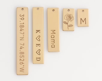 Puede incluir: Cinco etiquetas rectangulares de metal con diferentes grabados. Las etiquetas son de color beige y tienen un agujero en la parte superior para colgar. Los grabados incluyen una rosa, las letras "M", "Mama", "K ♥ E ♥ D" y las coordenadas "39.1847°N 74.8526°W".