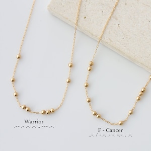 May include: Two gold necklaces with small round beads. The necklace on the left has the word "Warrior" written below it. The necklace on the right has the text "F - Cancer" written below it.