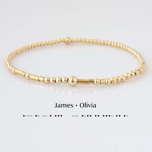May include: A gold beaded bracelet featuring a Morse code pattern. The bracelet consists of small round gold beads and elongated gold cylindrical beads. Below the bracelet, the text James • Olivia is printed with the corresponding Morse code dots and dashes.