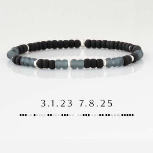 May include: A beaded bracelet featuring black and gray beads with silver accents. The bracelet has a simple design and is strung on an elastic cord. The image also includes the numbers "3.1.23 7.8.25" and their corresponding Morse code.