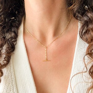 May include: A gold chain necklace with a long, thin pendant that has a horizontal bar at the bottom.