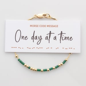 May include: Gold bracelet with green beads and a clasp. The bracelet has a card attached to it that says "Morse Code Message" and "One day at a time".