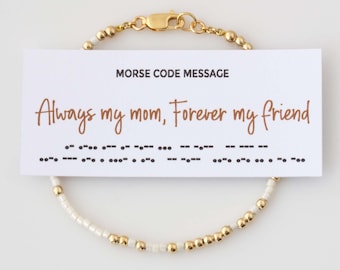 Könnte beinhalten: Ein zartes Armband mit abwechselnden goldenen und weißen Perlen und einem Karabinerverschluss. Eine weiße Karte liegt auf dem Armband und zeigt den Text "MORSE CODE MESSAGE" sowie "Always my mom, Forever my friend" mit Morsezeichen.