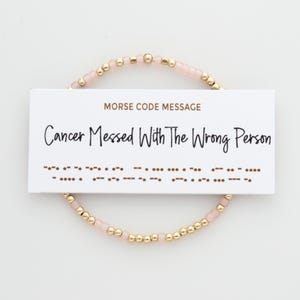 May include: A pink and gold beaded bracelet with a card that reads "Morse Code Message: Cancer Messed With The Wrong Person".