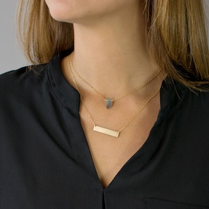 Op de afbeelding: Twee gouden kettingen met een klein, donkerblauw edelsteen hanger aan de ene en een dunne, rechthoekige staaf hanger aan de andere.