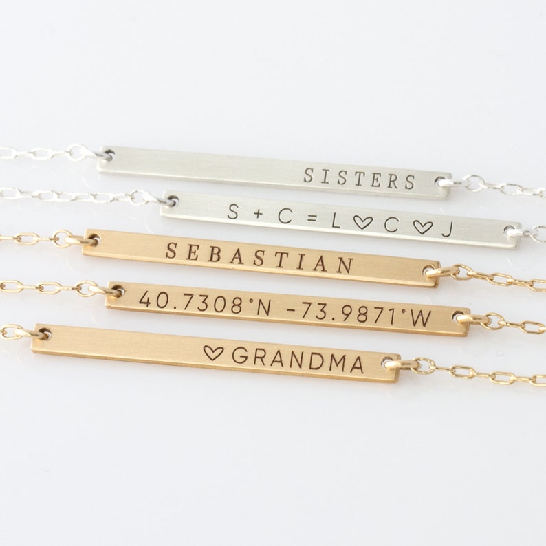 Puede incluir: Cuatro collares de barra de oro y plata con diferentes textos grabados. El collar superior dice "SISTERS". El segundo collar dice "S+C=L♡C♡J". El tercer collar dice "SEBASTIAN". El cuarto collar dice "40.7308&deg;N -73.9871&deg;W" y "♡GRANDMA".